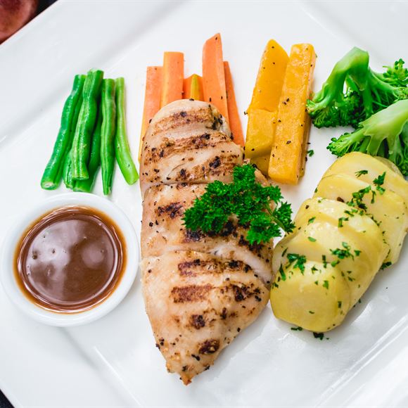 A grilled chicken breast plated with vegetables and a jug of sauce.