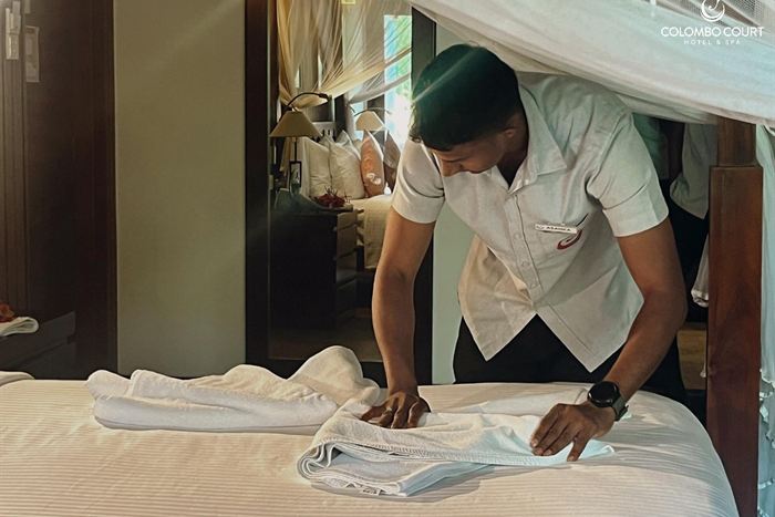 A member of the housekeeping team smoothing a white sheet across a bed under a four-poster frame.