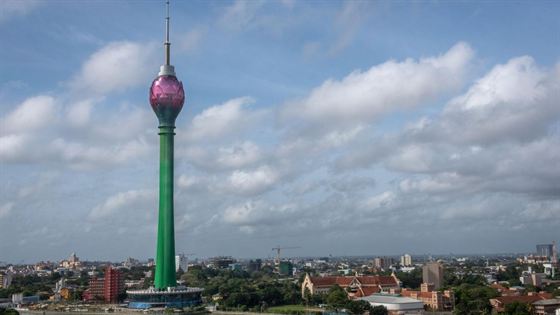 The Lotus Tower and the Colombo skyline under a cloudy sky, low rooftops and trees in the foreground.