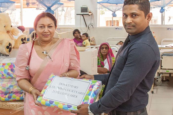 Two members of the Colombo Court team presenting a parcel of linen in a hospital ward, patients in the beds behind them.