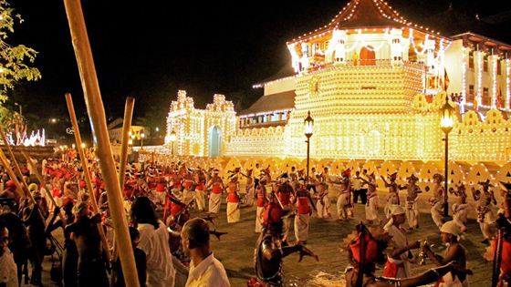 Kandyan drummers and dancers in white and red processing past the illuminated Temple of the Tooth at night.
