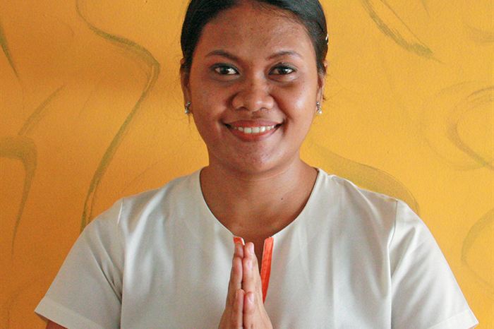 A therapist at The Court Spa in a white uniform, palms together in greeting, in front of a yellow wall.