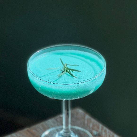 A pale blue cocktail served in a coupe glass, herb garnish across the top.
