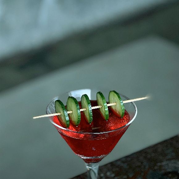 A deep red cocktail in a martini glass, cucumber slices on a skewer across the rim.
