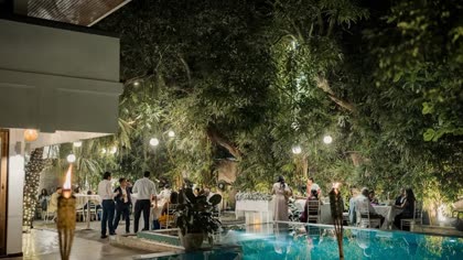 An evening reception in the poolside garden, guests standing with drinks beside the lit pool.