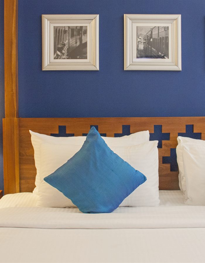 The head of a bed at Colombo Court, white linen and a blue cushion against a timber headboard and a deep blue wall.