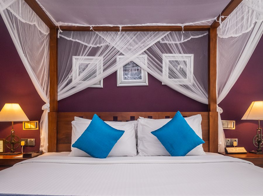 A Luxury Junior Suite at Colombo Court with a four-poster bed, draped net and a deep plum wall.